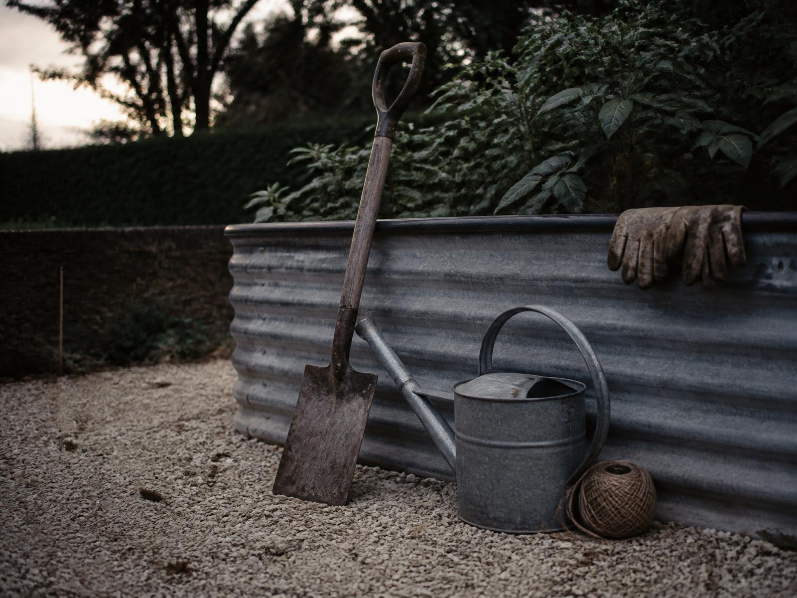 A spade, watering can, twine, and worn gloves left against a galvanized raised bed at dusk.
